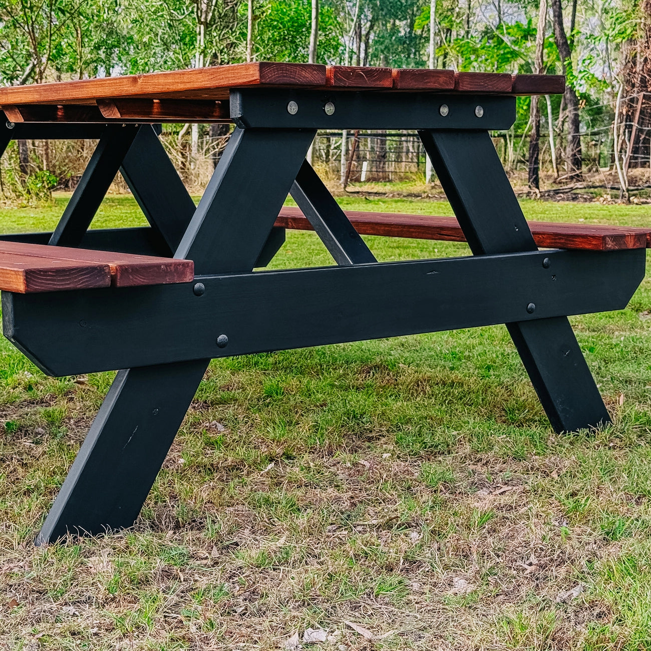 A black and brown wooden picnic table shown outdoors on a grassy lawn with trees in the background. Handmade by JJ Timber Design