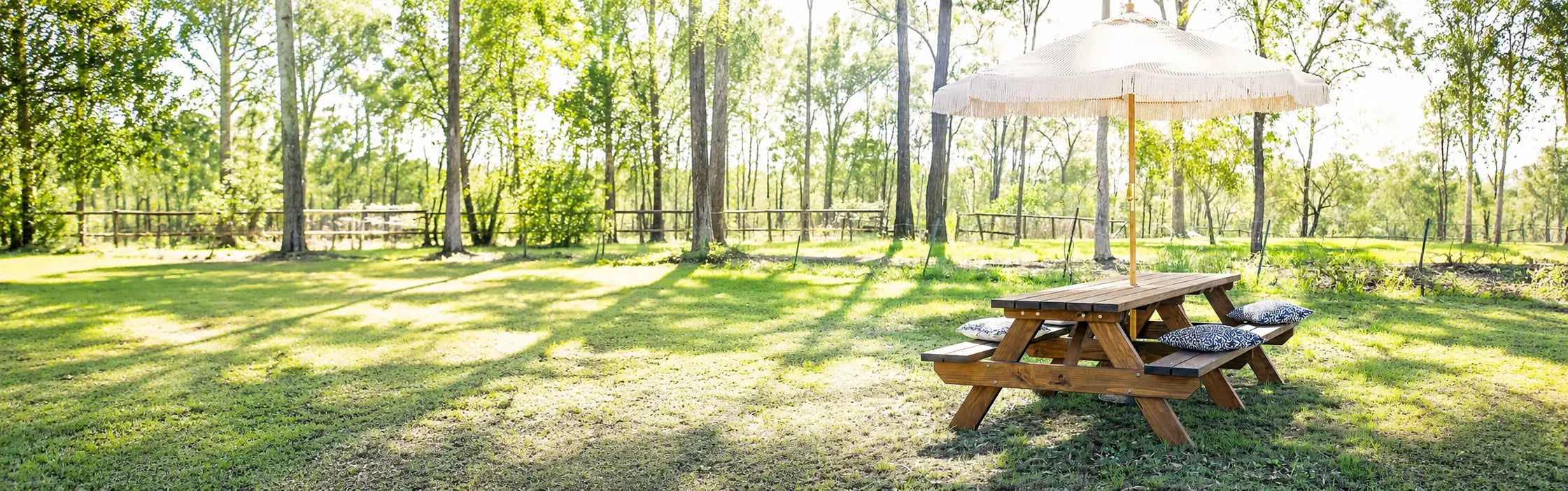 A light natural wood picnic table with attached benches sits on a bright green grassy lawn in a park-like setting with trees casting long  shadows.  JJ Timber Design Hero Banner
