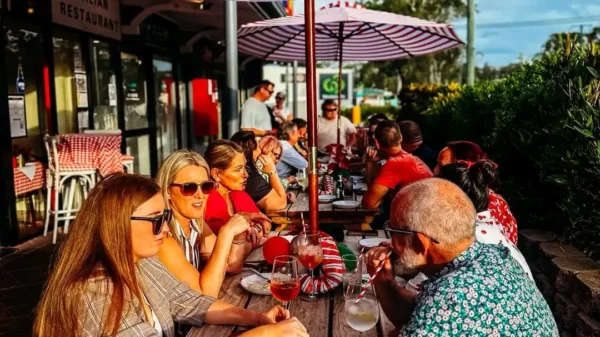 JJ Timber Design outdoor timber picnic table at Italian restaurant patio with people enjoying alfresco dining under striped umbrellas, premium hospitality furniture
